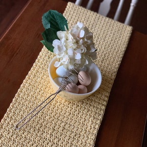 May include: A yellow crocheted table runner with a textured pattern. A bowl of eggs and a whisk sit on the runner, along with a small bouquet of white flowers. The runner is on a dark wooden table.
