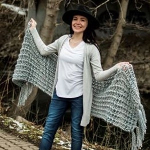 May include: A woman wearing a gray knitted shawl with fringe, a white t-shirt, blue jeans, and a black hat. She is standing on a brick path near a stream.