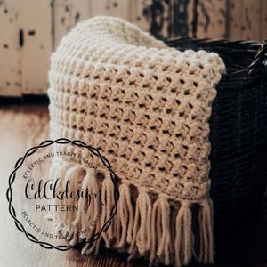 May include: A cream-colored crocheted throw blanket with fringe, draped over a dark wicker basket. The blanket has a textured, waffle-like stitch pattern.