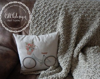 Könnte beinhalten: Eine beige gehäkelte Wolldecke mit einem strukturierten Muster liegt über einem braunen Ledersofa. Ein Kissen mit einem Blumendesign und einer Fahrrad-Illustration befindet sich auf dem Sofa.
