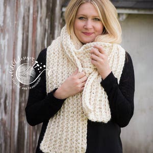 May include: A white crocheted scarf with a textured stitch pattern. The scarf is worn by a person with long blonde hair and a black shirt.