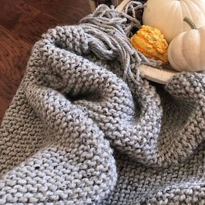 May include: A soft, grey knitted throw blanket with a fringe border. The blanket is draped over a white surface with decorative gourds and pumpkins.