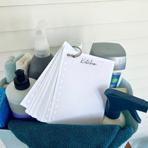 Op de afbeelding: Een blauwe plastic bak gevuld met schoonmaakmiddelen, waaronder een spuitfles, een spons, een handdoek en een notitieblok met de tekst "Keuken".