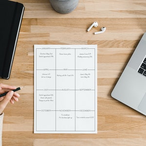 May include: A white paper calendar with black lines and text, detailing appointments and events. A black notebook, a laptop, and wireless earbuds are on a wooden surface. A hand holds a pencil.