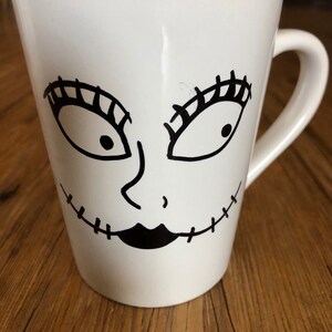 Puede incluir: Taza de cer&aacute;mica blanca con un dise&ntilde;o de cara de dibujos animados en negro. La cara tiene ojos grandes con largas pesta&ntilde;as, una nariz peque&ntilde;a y una sonrisa con la boca cosida. La taza tiene un asa curva y una forma ligeramente acampanada.