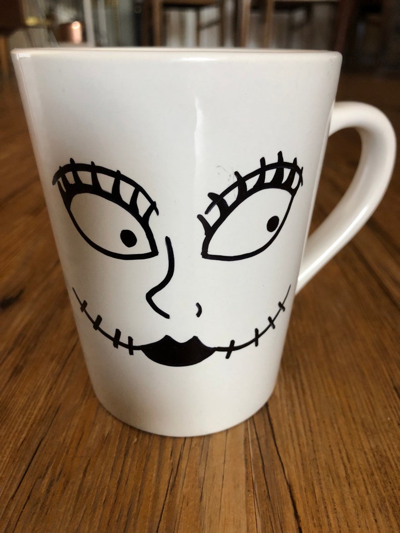 Puede incluir: Taza de cer&aacute;mica blanca con un dise&ntilde;o de cara de dibujos animados en negro. La cara tiene ojos grandes con largas pesta&ntilde;as, una nariz peque&ntilde;a y una sonrisa con la boca cosida. La taza tiene un asa curva y una forma ligeramente acampanada.