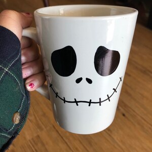 Puede incluir: Taza de cer&aacute;mica blanca con un dise&ntilde;o de rostro negro estilizado con ojos ovalados grandes y una sonrisa cosida. La taza tiene un asa y contiene una bebida. El dise&ntilde;o recuerda a un personaje animado popular.