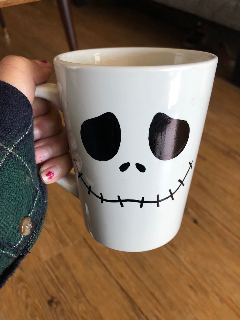 Puede incluir: Taza de cer&aacute;mica blanca con un dise&ntilde;o de rostro negro estilizado con ojos ovalados grandes y una sonrisa cosida. La taza tiene un asa y contiene una bebida. El dise&ntilde;o recuerda a un personaje animado popular.