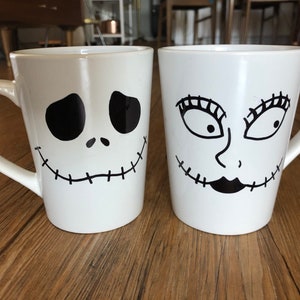 Puede incluir: Dos tazas de cer&aacute;mica blanca con dise&ntilde;os faciales negros. Una taza presenta una cara de Jack Skellington, la otra una cara de Sally, ambas de The Nightmare Before Christmas. Cada taza tiene un asa curva.