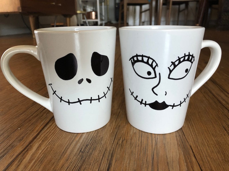 Puede incluir: Dos tazas de cer&aacute;mica blanca con dise&ntilde;os faciales negros. Una taza presenta una cara de Jack Skellington, la otra una cara de Sally, ambas de The Nightmare Before Christmas. Cada taza tiene un asa curva.