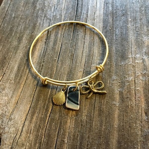 May include: Gold-colored adjustable bangle bracelet with three charms. Charms include a seashell, a rectangular piece with blue and white stripes, and a rope knot. The bracelet is displayed on a weathered wooden surface.