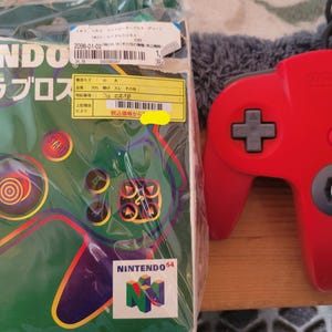 Peut inclure: Une manette rouge Nintendo 64 avec un pavé directionnel gris et des boutons colorés. À côté, une boîte verte avec du texte japonais et un graphique de manette. La boîte est emballée dans du plastique.