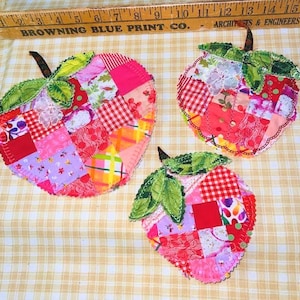 Könnte beinhalten: Drei apfelförmige Topflappen aus Stoff, gefertigt aus einem Patchwork verschiedener roter, rosa und bunter gemusterter Baumwollquadrate. Jedes Stück hat ein grünes Stoffblatt und einen braunen Stiel, mit einem sichtbaren Holzlineal oben.