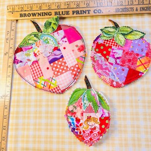 May include: Three fabric apple-shaped ornaments made of patchwork squares in pink, red, and green patterns with green fabric leaves. A wooden ruler shows the largest apple is approximately 5.5 inches wide and 6 inches tall.