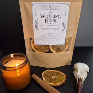 May include: A brown paper pouch labelled Lady Lunas Apothecary The Witching Hour Spiced Simmer Pot containing dried orange slices, with a lit candle in an amber glass jar, a cinnamon stick, a dried orange slice, and a bird skull nearby.