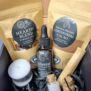May include: Aromatherapy gift set with a selection of items. The box contains two brown paper bags, a bottle of Ethereal bath oil, a small candle, two small glass jars, and a wooden stick. The bags are labelled "Hearth Blend" and "Ceremonial Cacao."