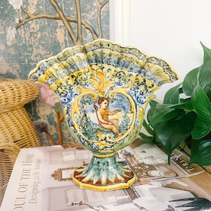 May include: A yellow, blue, and green ceramic vase with a scalloped rim and a cherub design. The vase is decorated with intricate floral patterns and has a green and gold base.