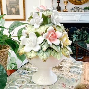 May include: A white ceramic floral arrangement featuring white, pink, and yellow flowers with green leaves. The flowers are arranged in a decorative vase with a pedestal base. The vase is white and has a glossy finish.