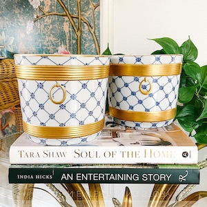 May include: Two white ceramic planters with blue and gold designs. The planters have a gold rim and a gold ring handle. The planters are sitting on top of two books. The top book is titled "Tara Shaw Soul of the Home". The bottom book is titled "India Hicks An Entertaining Story".