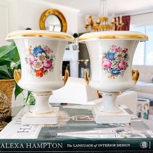 May include: Pair of white porcelain urns with gold trim and handles. Each urn features a floral bouquet in shades of pink, blue, and red. The urns are displayed on a table with a book titled "ALEXA HAMPTON The LANGUAGE of INTERIOR DESIGN."