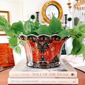 Può includere: Un vaso decorativo in ceramica con un design rosso e nero, pieno di piante verdi. Il vaso è appoggiato su due libri, "Soul of the Home" e "The Well Adorned Home."