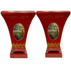 May include: A pair of red painted wooden plant stands with gold accents and a painted landscape scene. Each stand has a square base with three feet.