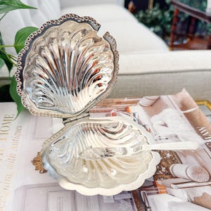 May include: A silver-colored, shell-shaped trinket box with an attached lid and a small spoon. The box is open, revealing the interior. The edges of the box and lid are decorated with a beaded design. The box is sitting on a magazine.