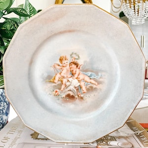 May include: A white porcelain plate with a gold rim features a hand-painted design of two cherubs with a dove. The cherubs are holding a wreath and a scroll.