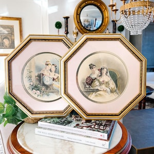 May include: Two framed vintage prints featuring women in period clothing. The frames are octagonal with gold trim and pink matting. The prints depict scenes with women in dresses and floral accents, evoking a classic aesthetic.