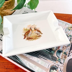 May include: A rectangular white ceramic ashtray with a raised border and two cigarette rests. The center features a detailed illustration of two brown and tan birds in a field. The ashtray is placed on a magazine.