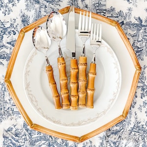 May include: A set of stainless steel silverware with bamboo-style handles, including a spoon, knife, and fork, arranged on a white plate with a gold rim. The plate is set on a patterned tablecloth with a blue and white design.