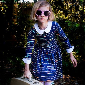 Peut inclure: Une jeune personne portant une robe rayée bleu et blanc avec un col blanc et des lunettes de soleil en forme de cœur rose. Elle tient une valise vintage de couleur brun clair.