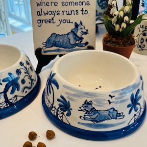 May include: White ceramic dog bowl with a blue painted design featuring palm trees, a running dog, and the phrase "where someone always runs to greet you." The bowl has a blue rim and is filled with dog food.