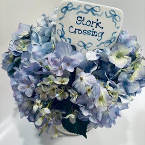 May include: A bouquet of blue hydrangeas with a white sign that says "Stork Crossing" in blue lettering.