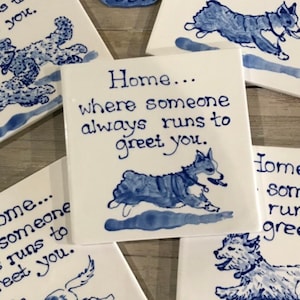 May include: A white ceramic tile with blue lettering and a blue illustration of a dog running. The text reads "Home... where someone always runs to greet you."
