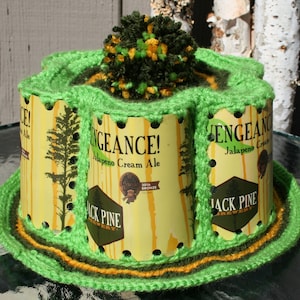 May include: A green crocheted hat with a green and yellow pom-pom on top. The hat is made from recycled beer can tabs and features the words "Vengeance! Jalapeno Cream Ale" and "Jack Pine Brewery".