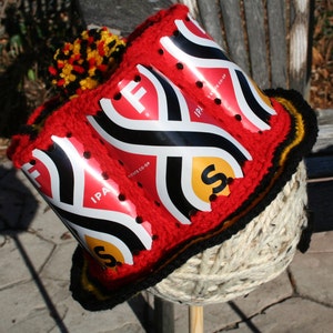 May include: A red crocheted hat with a black and yellow trim. The hat is made from recycled aluminum cans with the words "IPA" and "SIXTE CO-OP" printed on them. The hat is topped with a black, yellow, and red pom-pom.