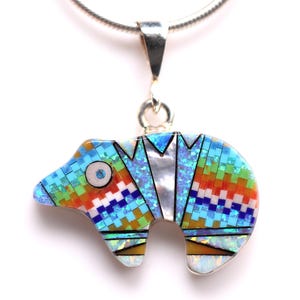 May include: A colorful bear pendant with a silver chain. The bear is made of inlaid stones in shades of blue, orange, green, and white. The pendant has a detailed design and is set against a white background.