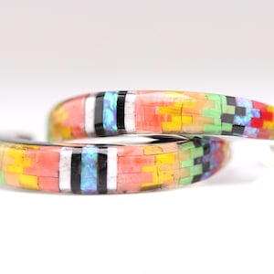 May include: A pair of hoop earrings featuring a mosaic inlay of small rectangular stones in vibrant shades of red, yellow, green, black, white, and iridescent blue. The stones are arranged in a geometric pattern around the curved surface.