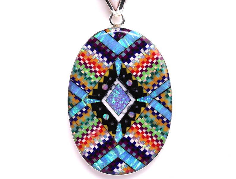 May include: A silver pendant with a colorful geometric design made of inlaid stones. The design features a central diamond shape with four points, surrounded by a border of smaller squares and rectangles. The stones are a variety of colors, including blue, green, red, orange, yellow, and black.