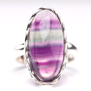 Puede incluir: Un anillo de plata con una gran piedra preciosa de fluorita de forma ovalada. La piedra preciosa es una mezcla vibrante de rayas moradas, rosas y blancas.