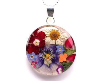 Op de afbeelding: Een ronde hangerketting met een heldere harskoepel met verschillende gedroogde bloemen. De bloemen zijn onder meer rode, witte, blauwe, paarse en oranje bloesems. De hanger hangt aan een zilveren ketting.