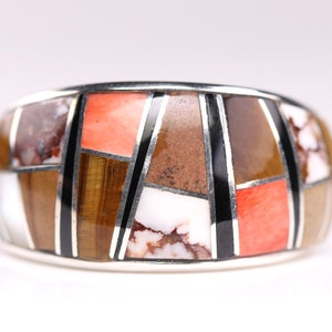 May include: A wide silver band ring with a geometric pattern of inlaid stones. The stones are in shades of brown, white, orange, and black.