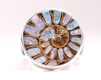 May include: A silver ring featuring a polished ammonite fossil. The fossil displays a spiral pattern with shades of brown, purple, and blue. The ring has a wide band and a circular setting.