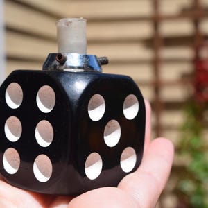 May include: A black dice-shaped perfume bottle with white dots. The bottle has a silver metal top and a clear, cylindrical stopper. The bottle is held in a hand against a blurred outdoor background.