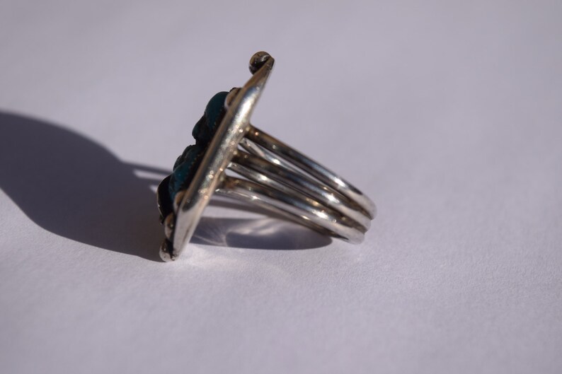 Puede incluir: Un anillo de plata con una piedra turquesa engastada en un dise&ntilde;o geom&eacute;trico. El anillo tiene una banda ancha con m&uacute;ltiples bandas.