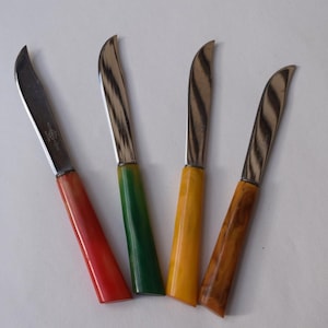 May include: Four cheese knives with curved blades and colorful handles. The handles are red, green, yellow, and brown. The blades have a striped pattern. The knives are arranged on a white surface.