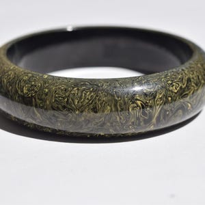 May include: A smooth, polished bangle bracelet featuring a dark, swirling pattern of black and metallic gold colors. The exterior surface displays an intricate, marbled design, while the interior of the circular band is a solid, glossy black finish.