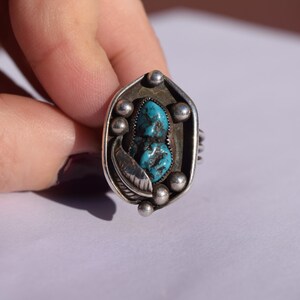 Puede incluir: Un anillo de plata con una piedra turquesa engastada en un dise&ntilde;o geom&eacute;trico. El anillo est&aacute; adornado con peque&ntilde;as cuentas de plata y un detalle en forma de pluma.