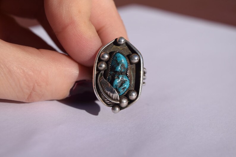 Puede incluir: Un anillo de plata con una piedra turquesa engastada en un dise&ntilde;o geom&eacute;trico. El anillo est&aacute; adornado con peque&ntilde;as cuentas de plata y un detalle en forma de pluma.